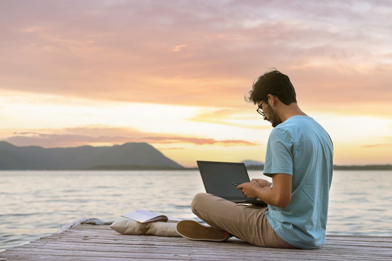 A man sits on a wooden dock by a lake, using a laptop at sunset. Mountains and calm water are in the background, and an open book—perhaps reflecting Baby Boomer beliefs about lifelong learning—lies beside him.