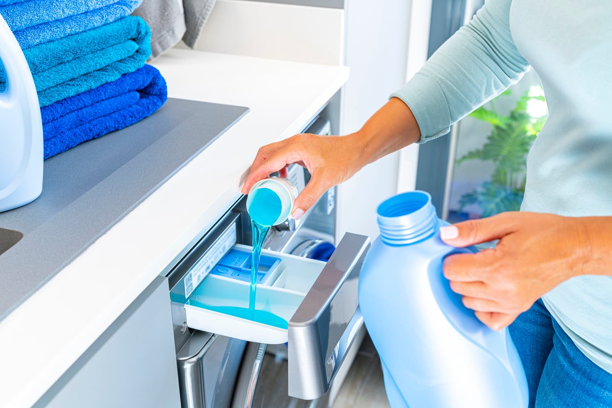 A person pours blue liquid laundry detergent—one of those products people no longer buy as frequently—into a washing machine drawer, with folded blue towels and detergent bottles nearby.