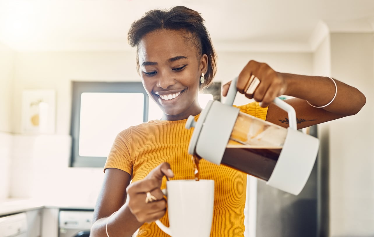A woman in an orange shirt smiles while pouring coffee from a French press into a white mug in a bright kitchen, embracing money saving habits by making her favorite brew at home.
