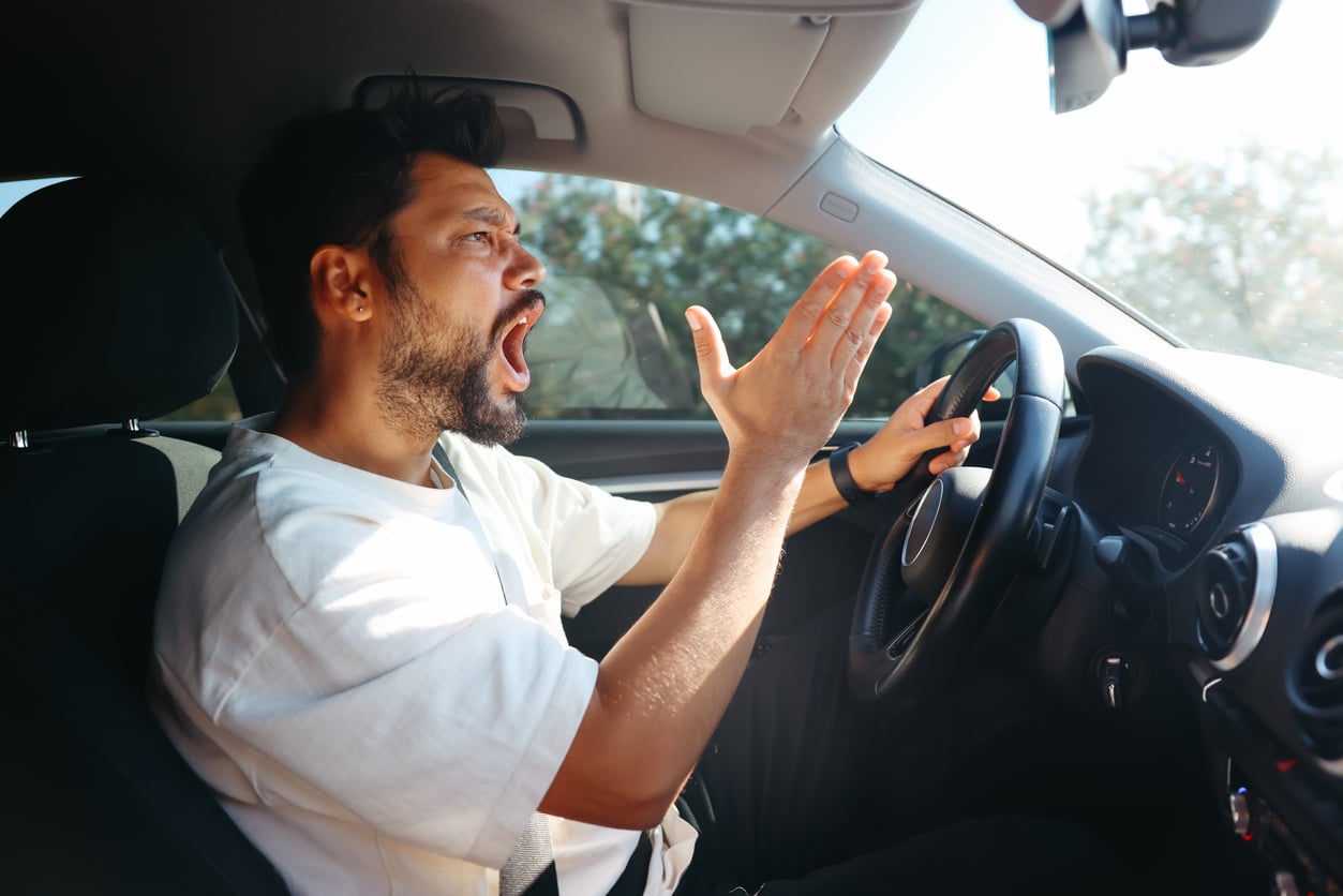 A man sits in the driver's seat of a car, yelling and gesturing angrily with his right hand while holding the wheel with his left—perhaps wishing he knew some car maintenance hacks to avoid on-road frustrations during the day.