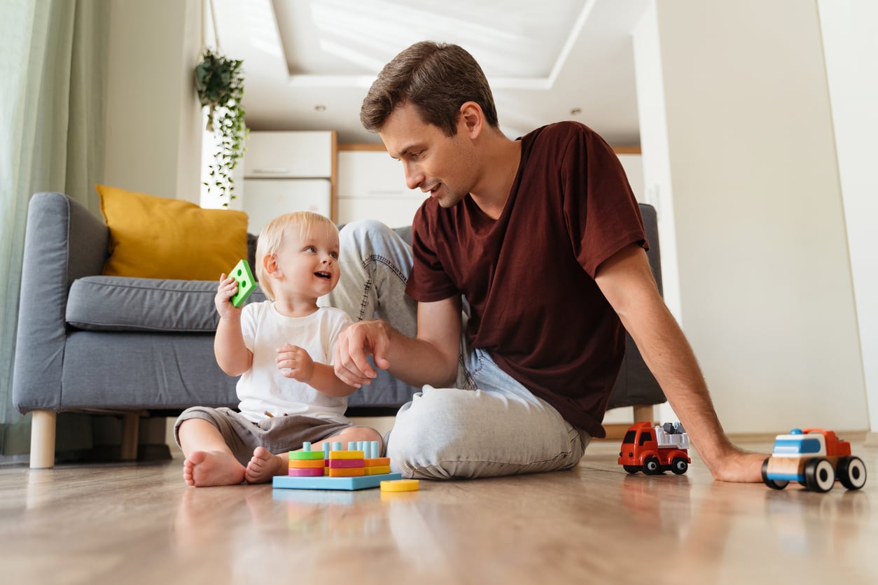 A smiling man and toddler sit on a living room floor, playing with colorful blocks and toy cars. Reflecting Baby Boomer beliefs in the value of quality time, the man encourages hands-on play as the toddler holds up a green block; both appear happy and engaged.