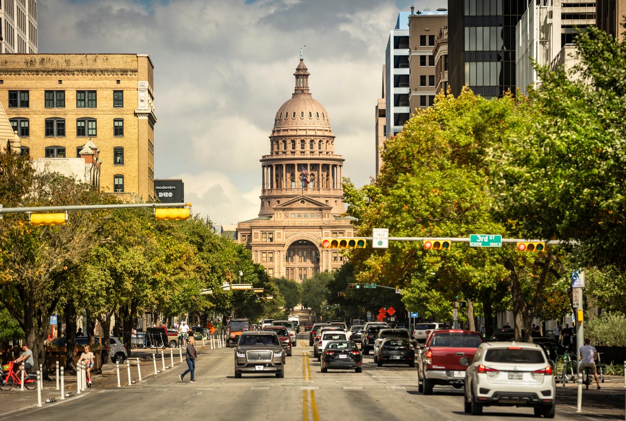 A busy city street lined with trees and cars leads to a large domed building, the Texas State Capitol, under a partly cloudy sky in Austin, Texas.