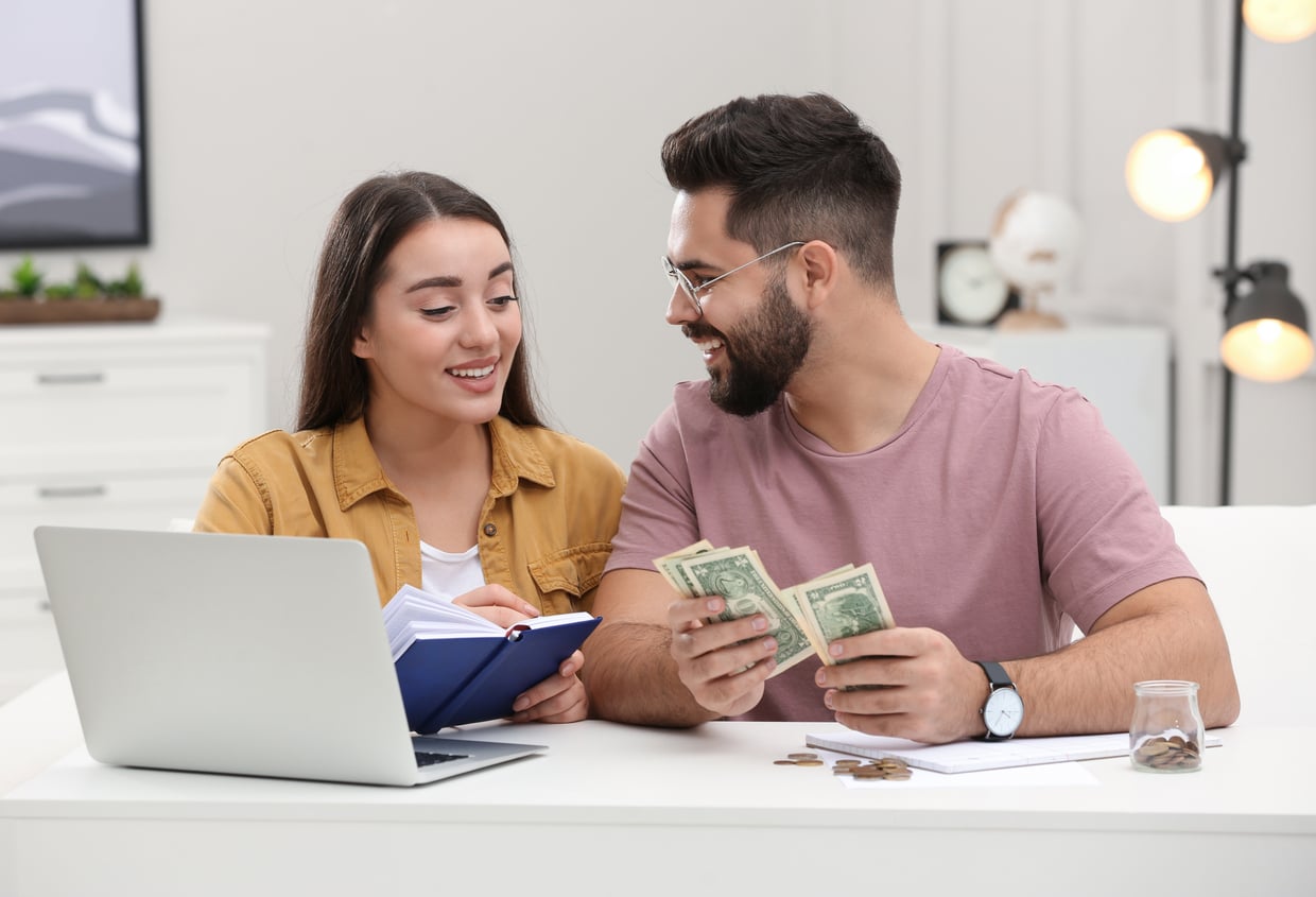 A smiling couple sits at a table with a laptop, notebook, and cash as they discuss the benefits of renting. The man counts dollar bills while the woman writes in a notebook; coins, a jar, and papers suggest they're budgeting together.