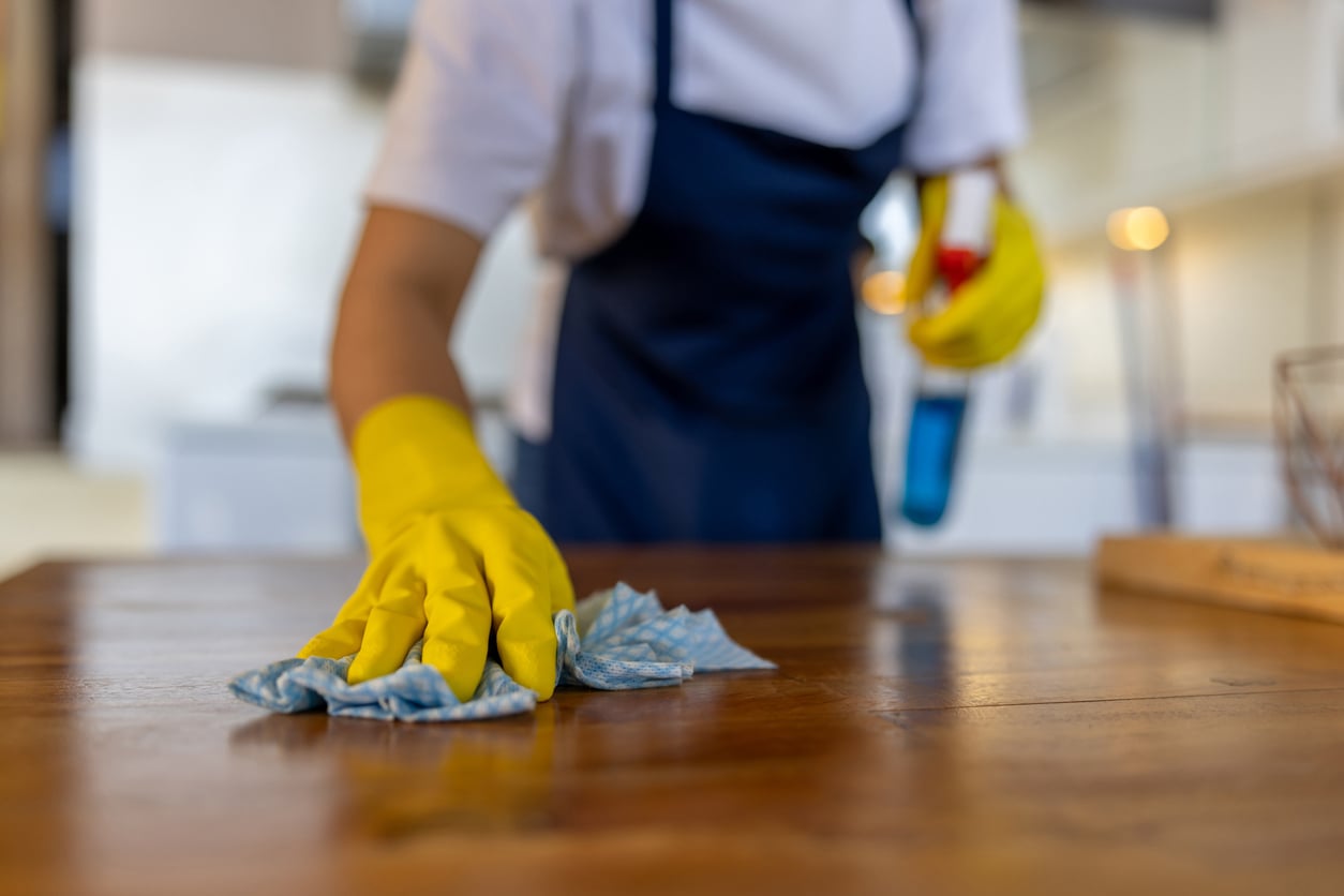 A person wearing yellow gloves wipes a wooden table with a blue and white cloth. A bottle of cleaning spray is in the background.