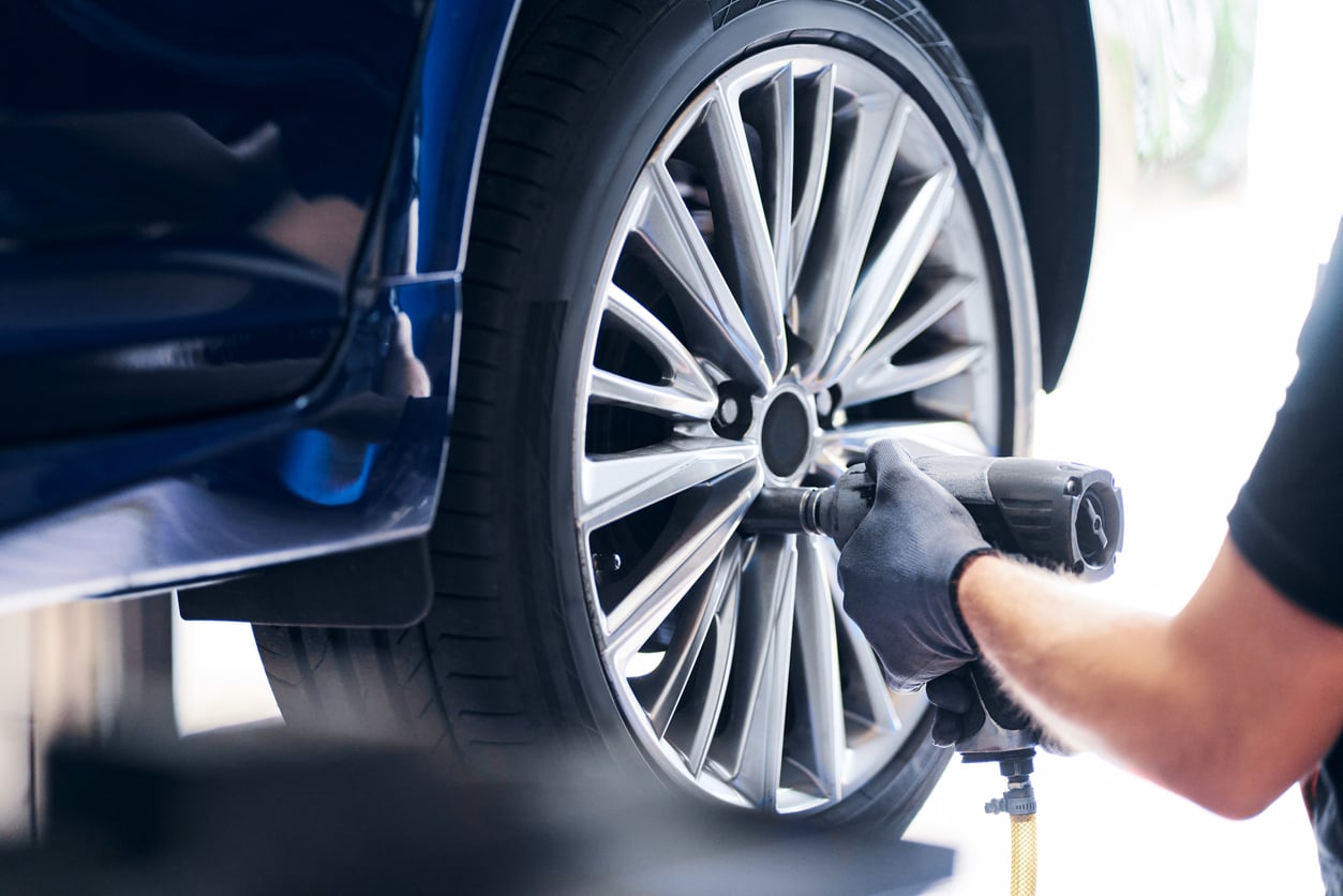 A person wearing black gloves uses products like a power tool to tighten or remove the lug nuts on a car's silver alloy wheel. The car appears to be lifted for maintenance or a tire change.