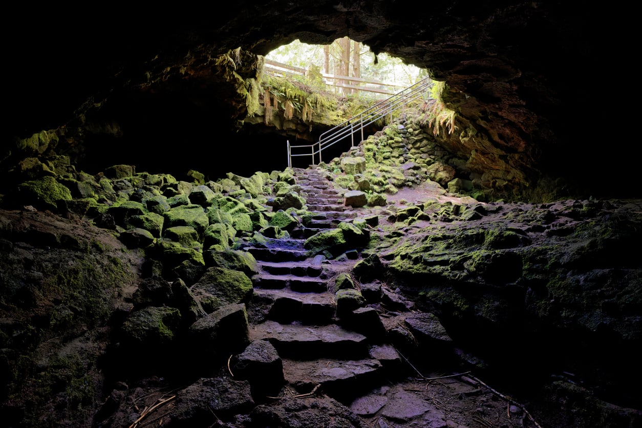 Stone steps lead out of a cave covered in moss and rocks, with sunlight shining through the entrance above. A metal railing lines the upper steps near the exit, surrounded by greenery and trees outside—making this hidden gem one of the captivating travel destinations on the West Coast.