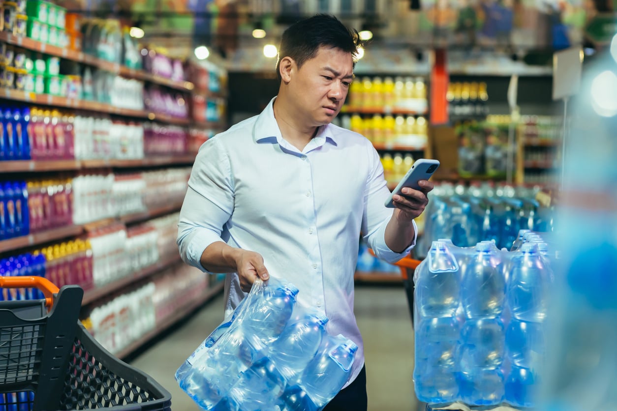A man in a white shirt holds bottled water packs in a supermarket aisle, looking at his phone with a concerned expression—perhaps questioning if bottled water is among the waste of money products. A shopping cart and shelves stocked with products are visible.
