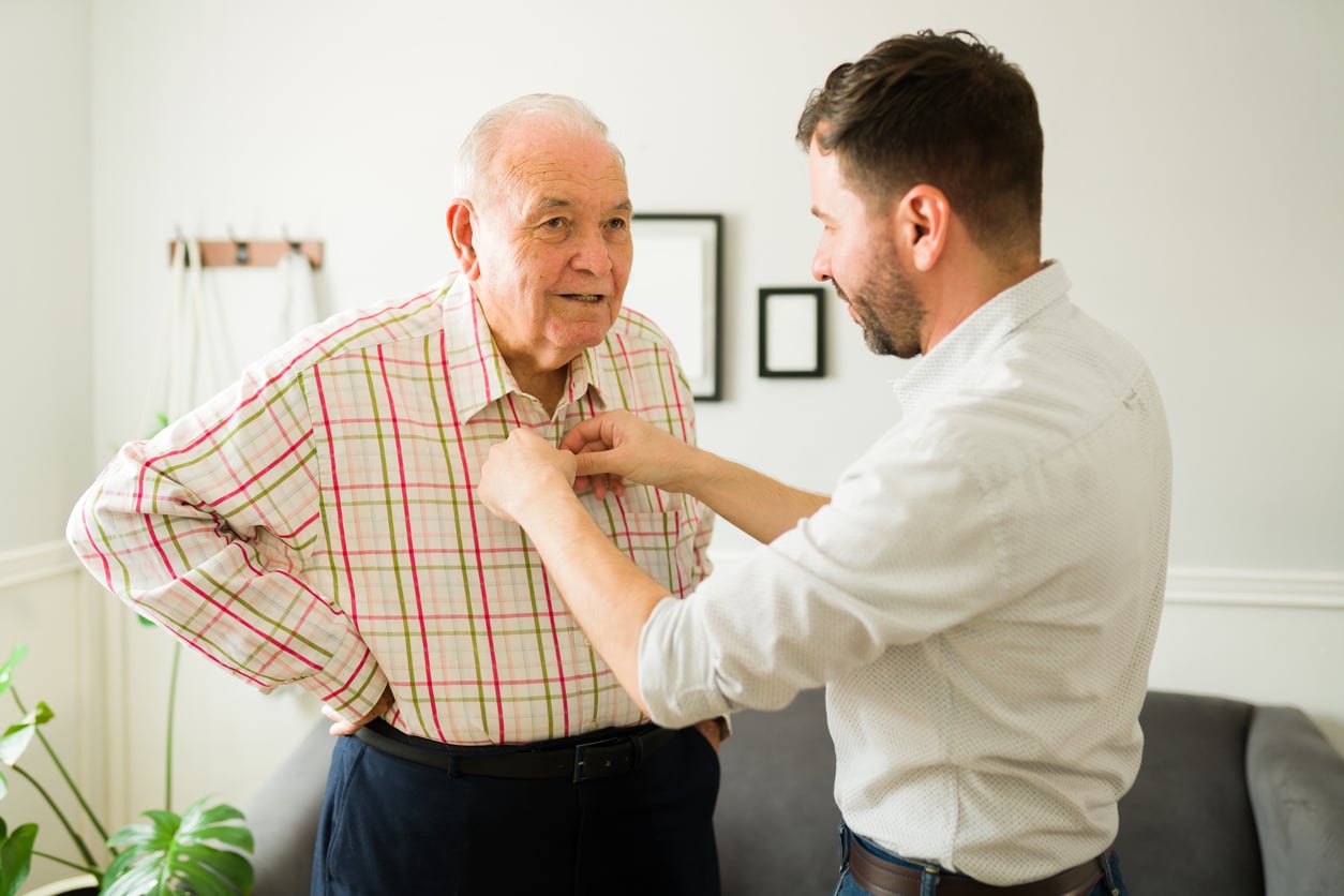 A younger man helps an older man button his plaid shirt while standing in a living room with neutral decor.