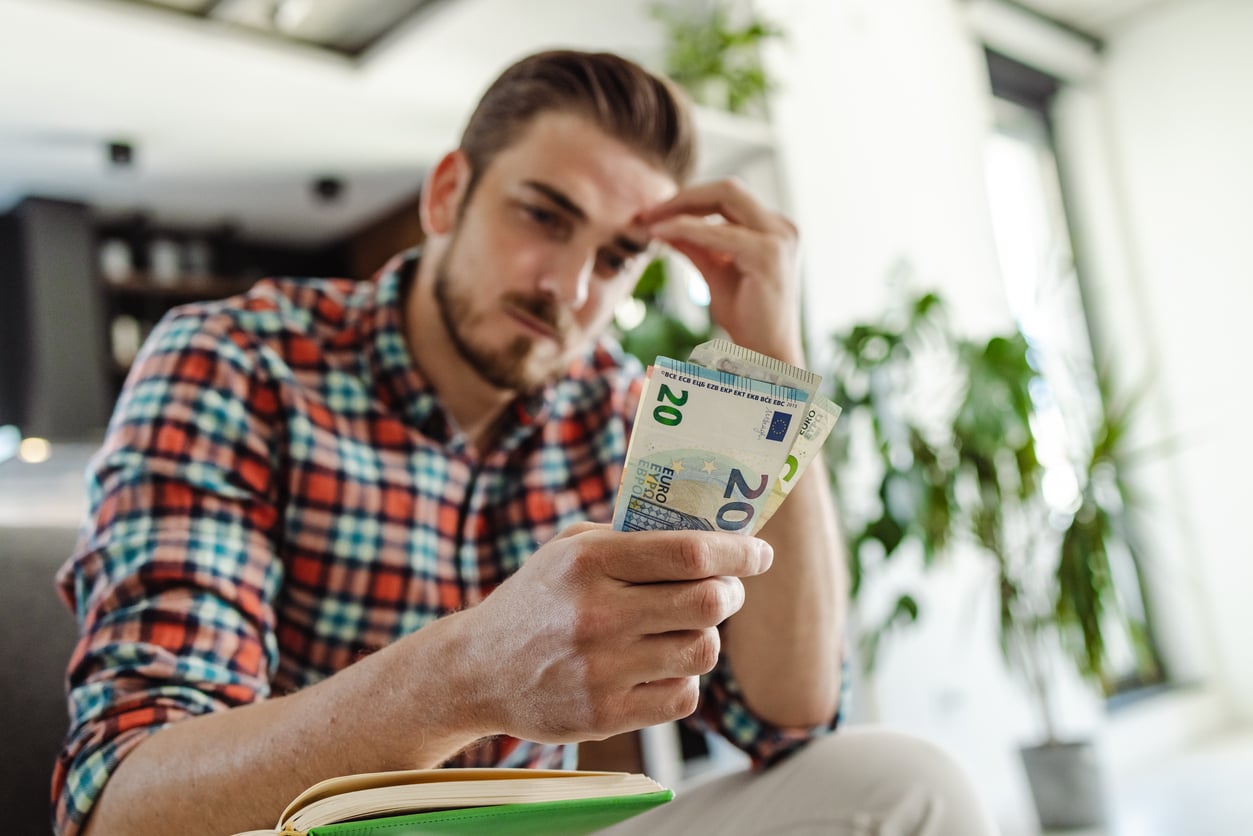 A man in a plaid shirt sits indoors, looking worried while counting euro banknotes with one hand and holding his head with the other. Green plants are visible in the background, suggesting he may be reflecting on his money saving habits.