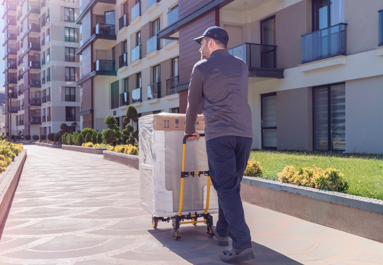 A delivery worker in uniform pushes a large, wrapped package—perhaps filled with products people no longer buy—on a trolley along a sunny sidewalk outside modern apartment buildings.
