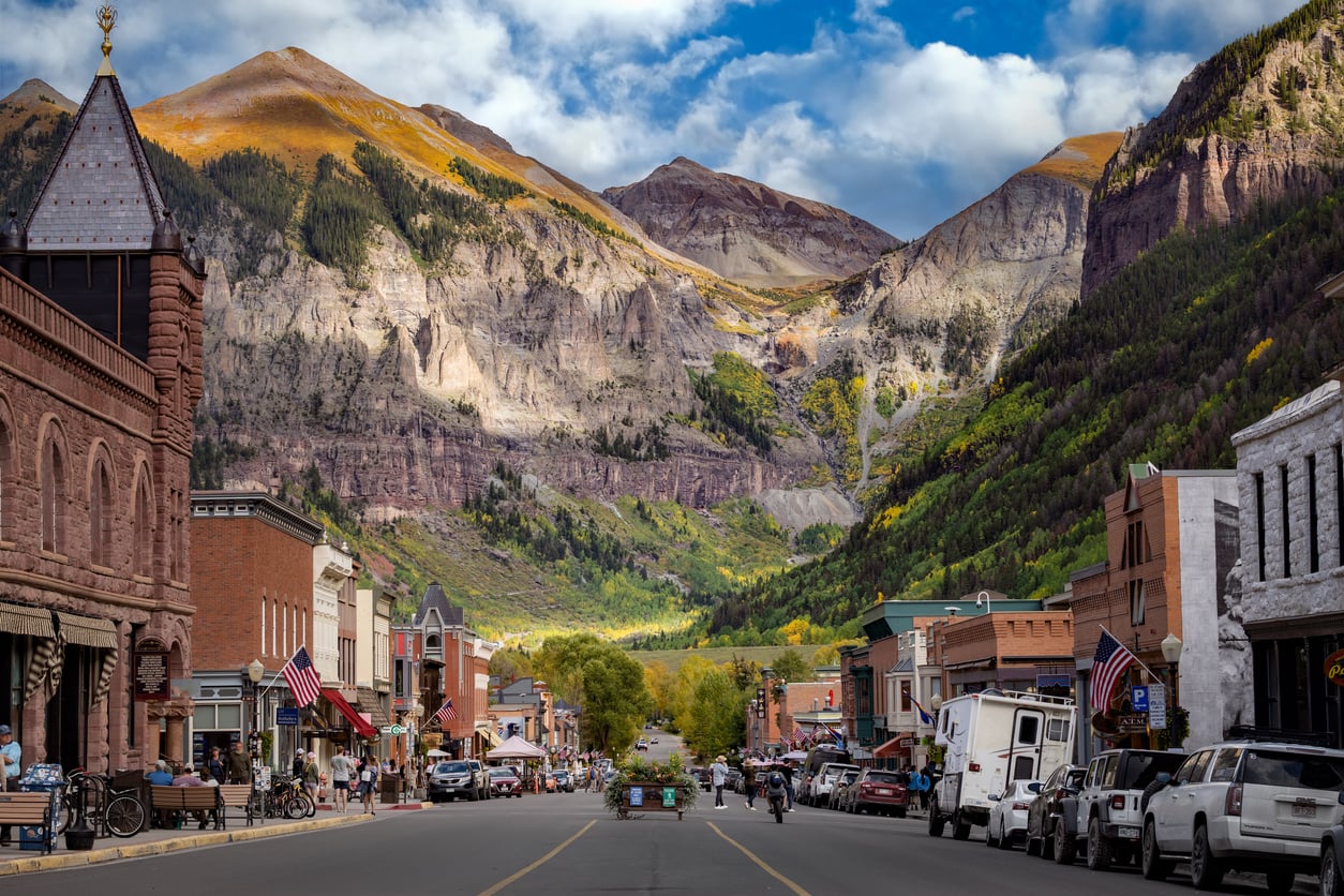 Small town street with brick buildings, American flags, and parked cars, set against dramatic mountains with patches of autumn foliage, under a partly cloudy sky—a picturesque scene that captures the charm of classic travel destinations.