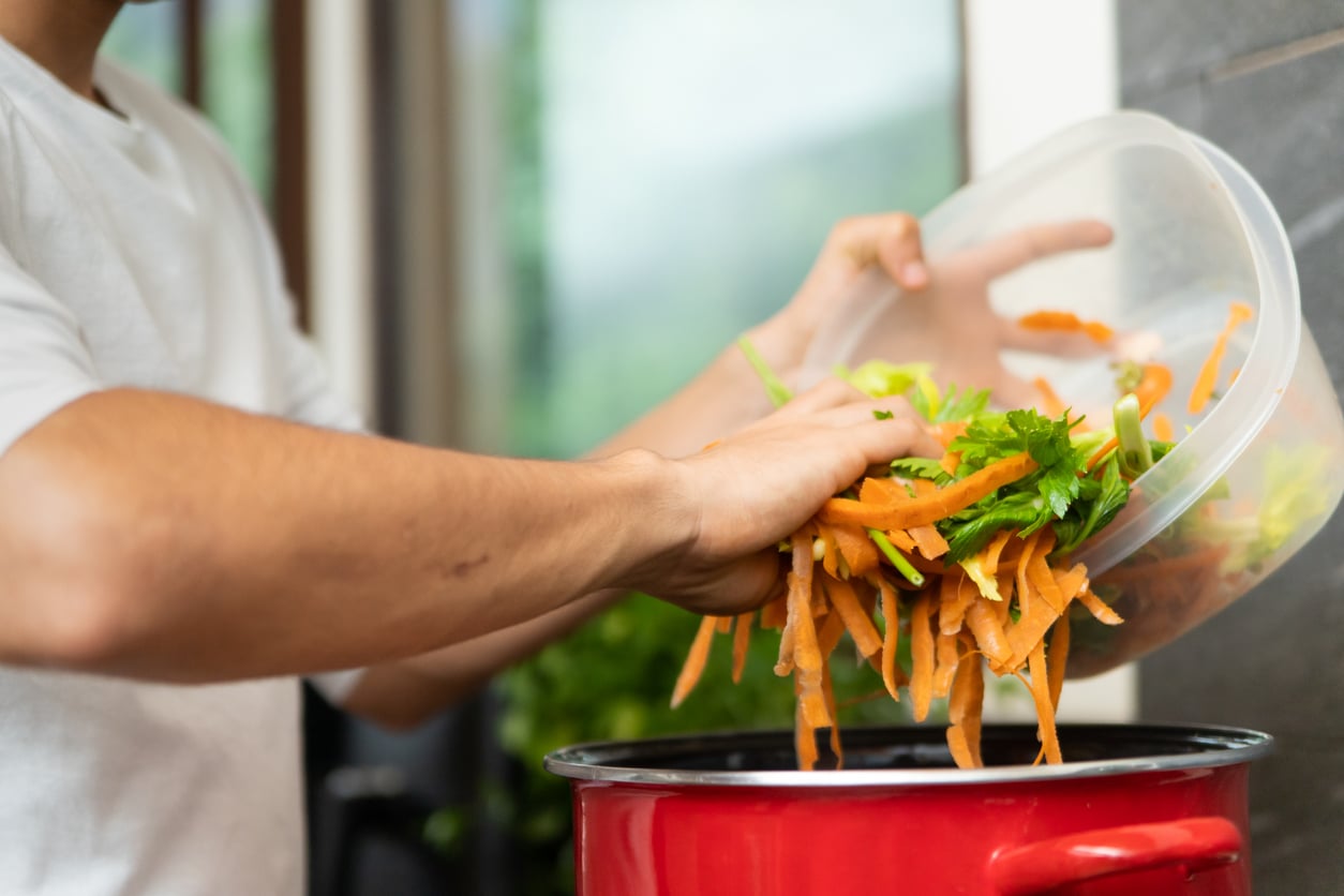 A person pours vegetable scraps, including carrot peels and parsley, from a container into a red pot, likely preparing to make vegetable stock in a kitchen—a simple way to cut down on monthly bills by making use of leftovers.