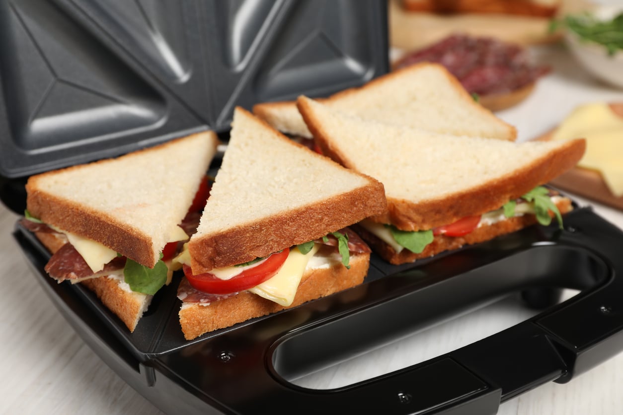 Four uncooked sandwiches with cheese, tomato, meat, and greens are placed inside a sandwich maker—one of the must-have kitchen gadgets—ready to be toasted. The sandwiches are cut diagonally and arranged in the appliance.