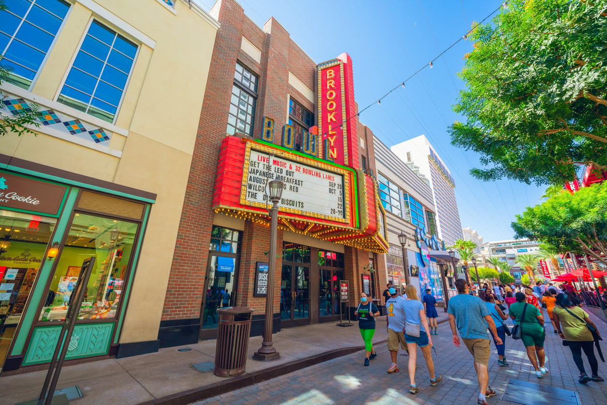 People walk along a sunny outdoor pedestrian street, passing by the Brooklyn Bowl and sharing movie theater hacks. Shops and trees line the street under a clear blue sky, with a colorful marquee sign listing live shows.