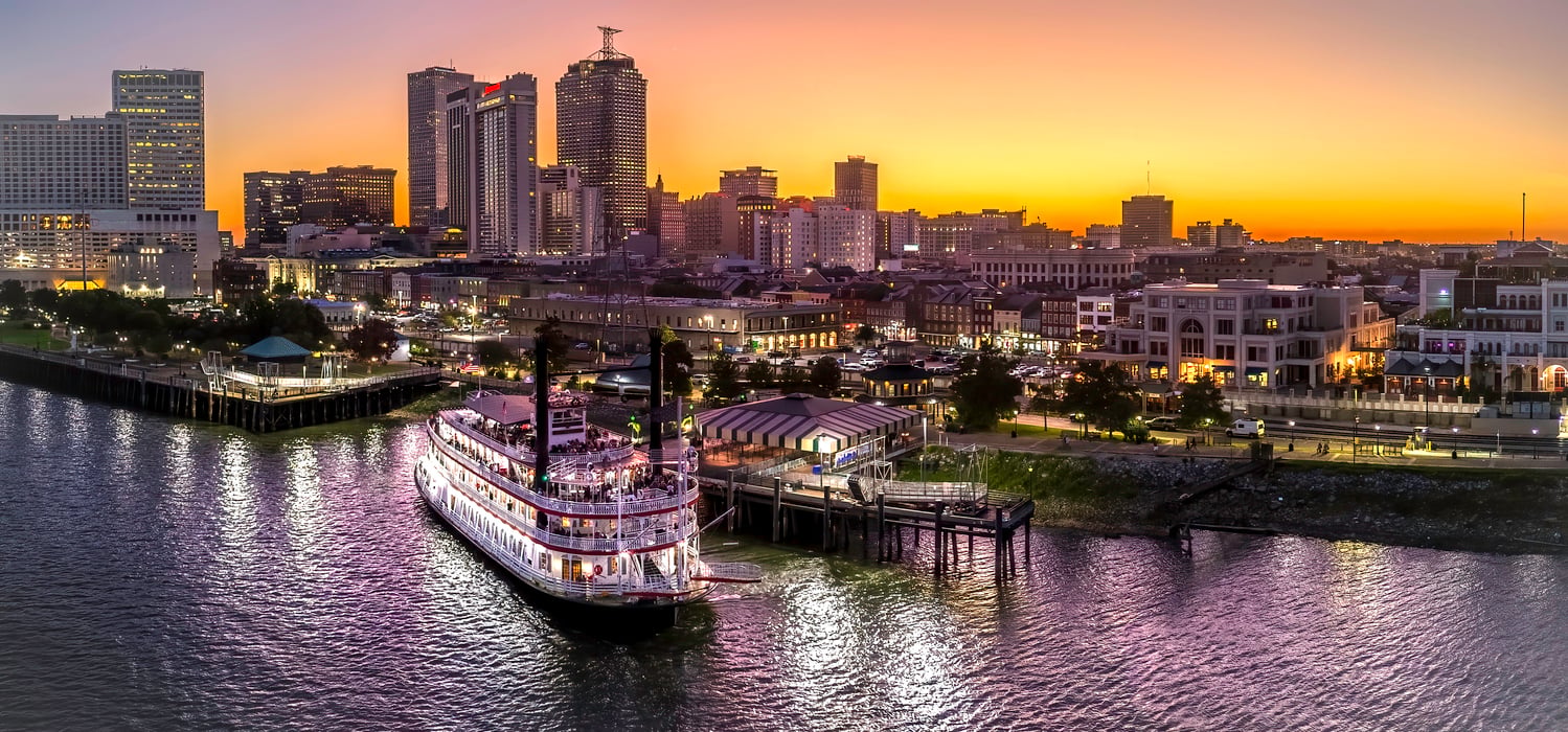 A paddlewheel riverboat docked along the waterfront in front of a lively city skyline at sunset, with vibrant reflections on the water and illuminated buildings in the background.