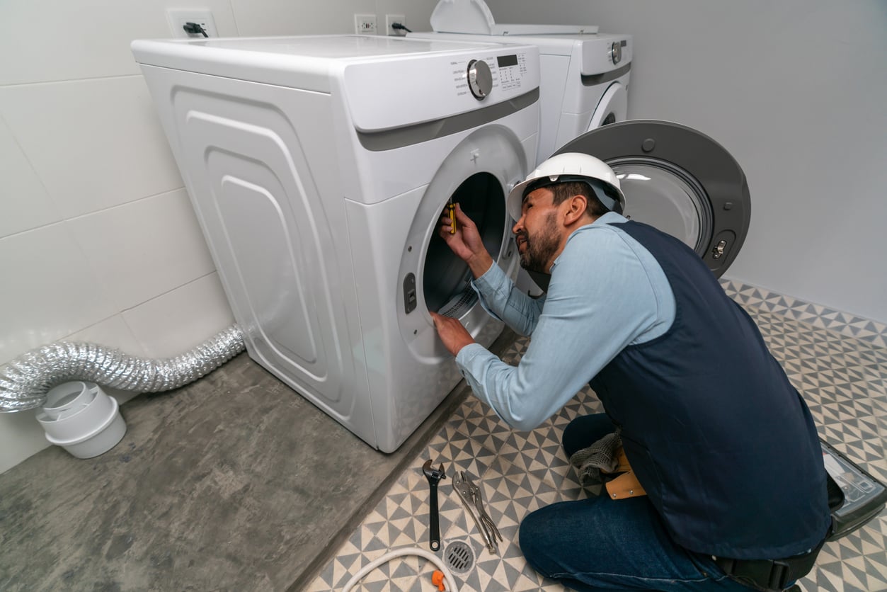 A repairman wearing a hard hat is kneeling and inspecting the inside of a front-loading washing machine in a laundry room, with tools, various products, and a dryer vent hose nearby.