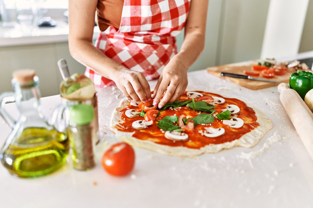 A person wearing a red checkered apron prepares homemade pizza, adding vegetables to dough topped with tomato sauce, mushrooms, and spinach. Olive oil, tomato, rolling pin, and spices are on the countertop.