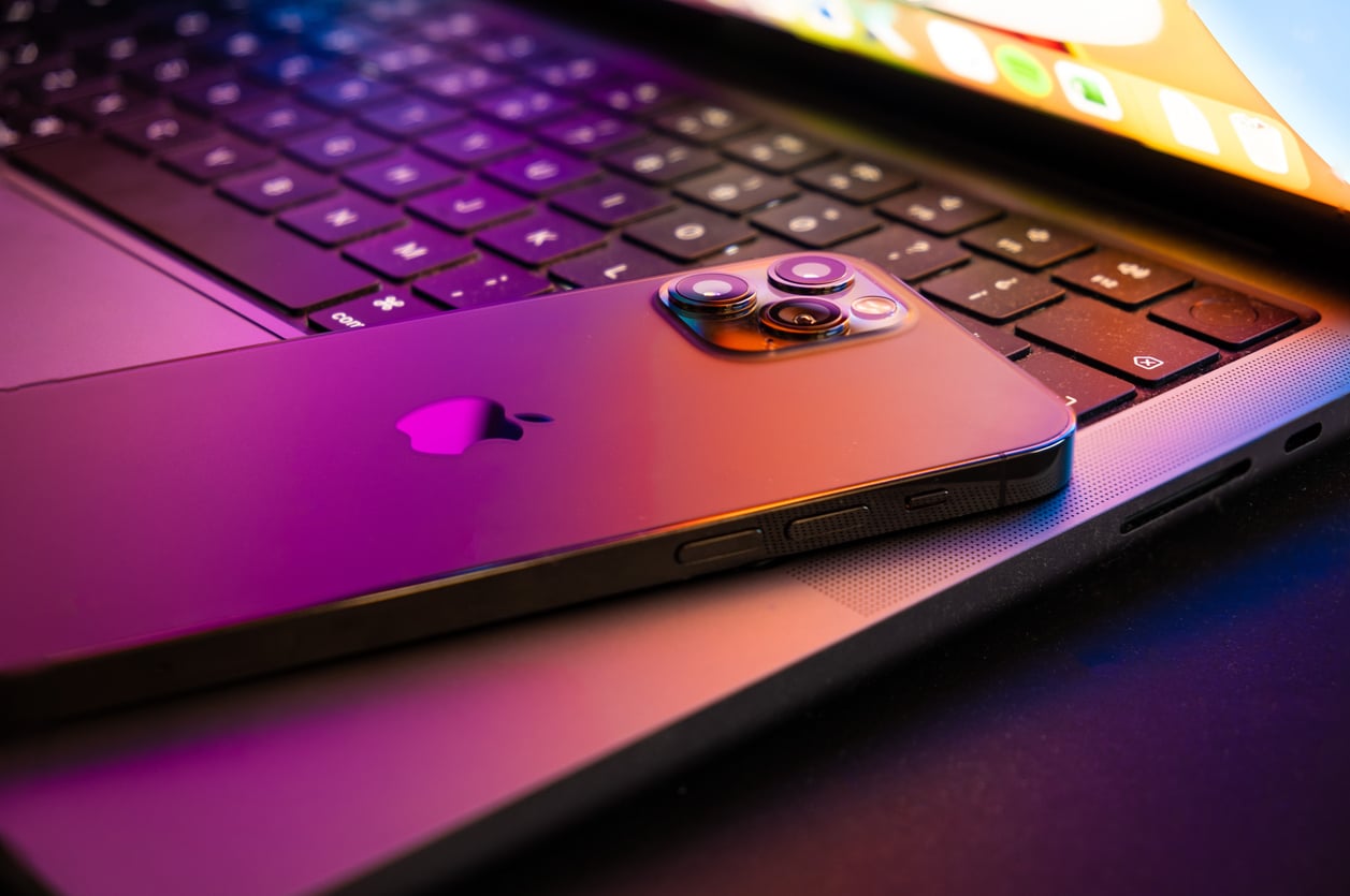A smartphone with a reflective Apple logo rests on the keyboard of an open laptop, both illuminated by colorful, ambient lighting.