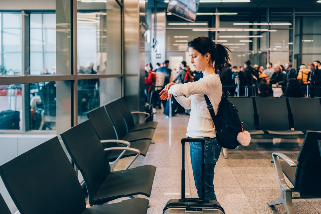 A woman with a backpack stands by her suitcase in an airport, checking her watch. Empty seats are around her, and a crowd of people is gathered in the background near the gate.