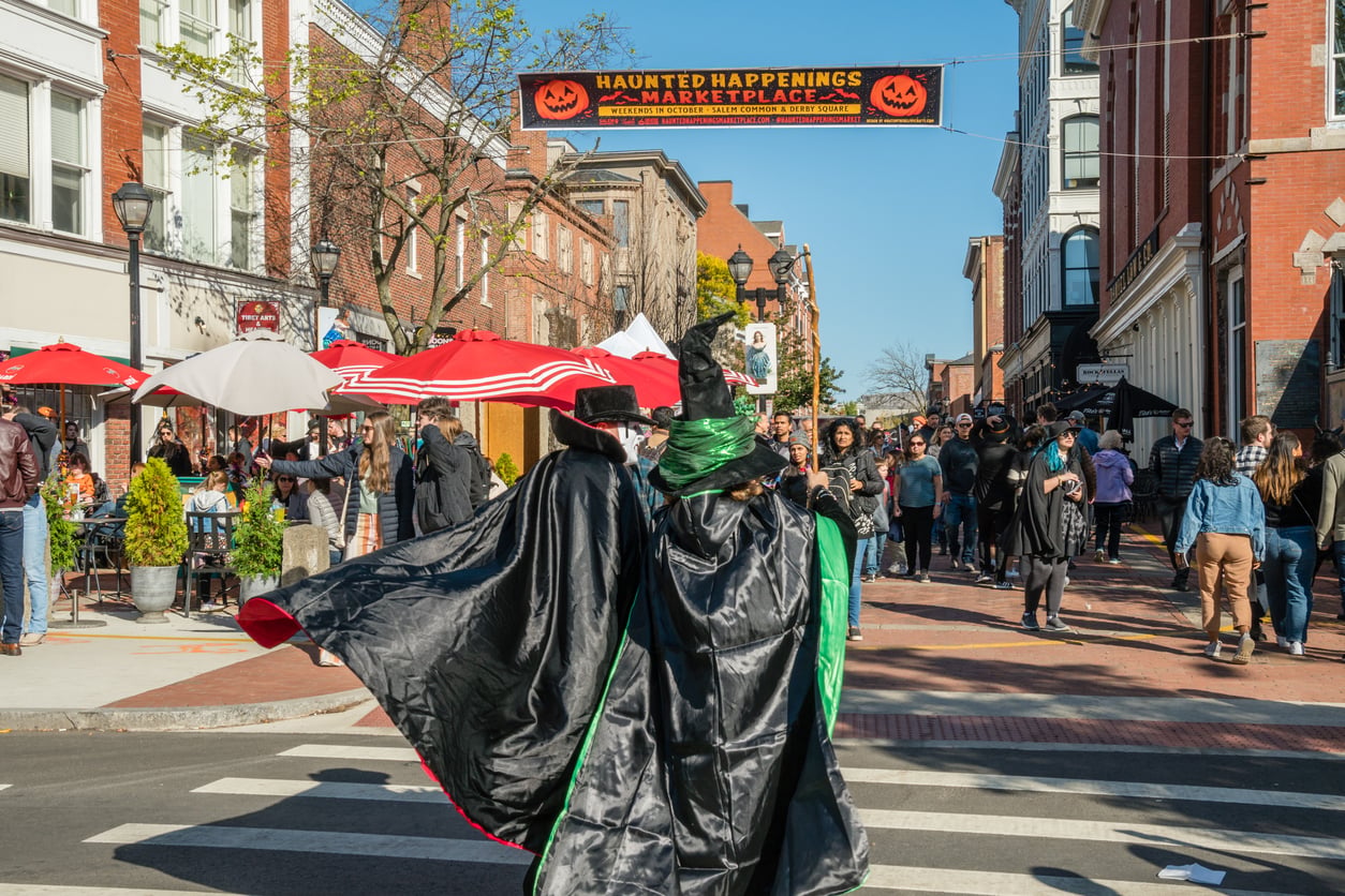 Two people in black and green witch costumes cross a street at a busy outdoor Halloween event, with a "Haunted Happenings Marketplace" sign and crowds under red umbrellas in the background.