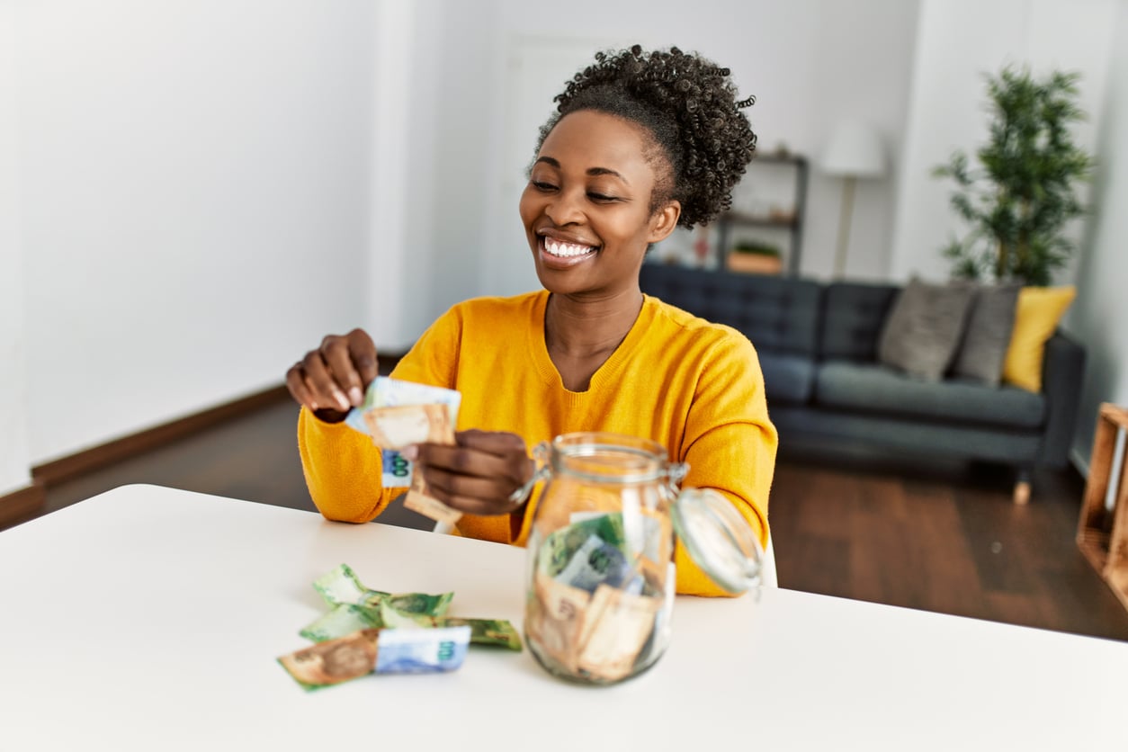 Smiling woman in a yellow sweater counts paper money at a table, with a glass jar full of cash in front of her, in a bright living room—showcasing how women are changing their spending behaviors.