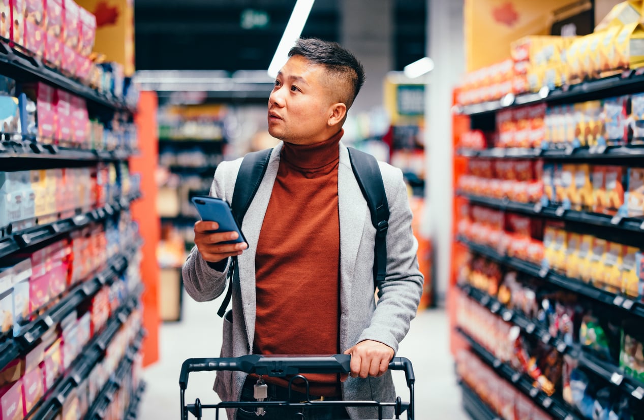 A person with a backpack pushes a shopping cart down a grocery store aisle, holding a smartphone and looking up at the shelves—clearly practicing money saving habits while making thoughtful choices.