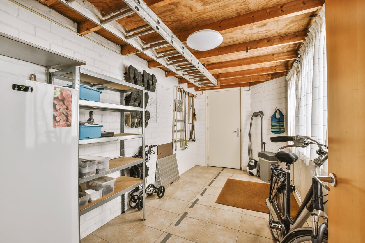 A tidy utility room with white brick walls, open wooden ceiling beams, a metal storage shelf, a bicycle, a refrigerator, cleaning supplies, and a window letting in natural light.