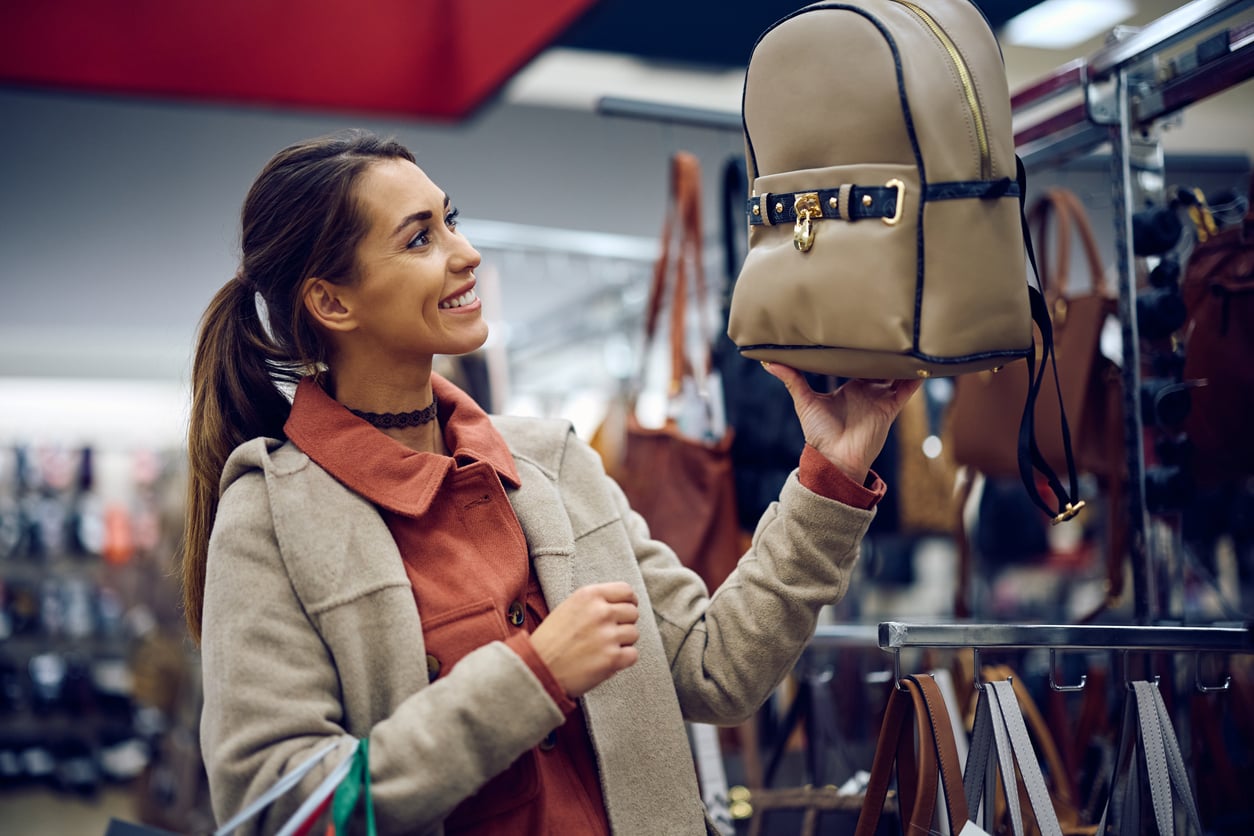 A woman in a beige coat smiles while holding up a tan backpack in a store, surrounded by various purses and bags on display racks.