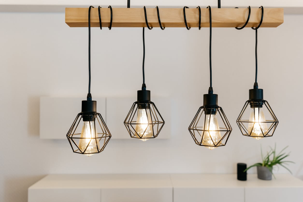 Four modern pendant lights with geometric wire shades hang from a wooden beam, glowing warmly in a minimal, contemporary room with white walls and simple decor.