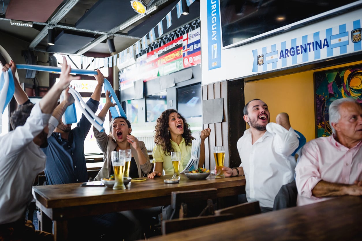 A group of people in a bar, cheering and celebrating while watching a sports event, with drinks and snacks on the table and an Argentina banner on the wall.