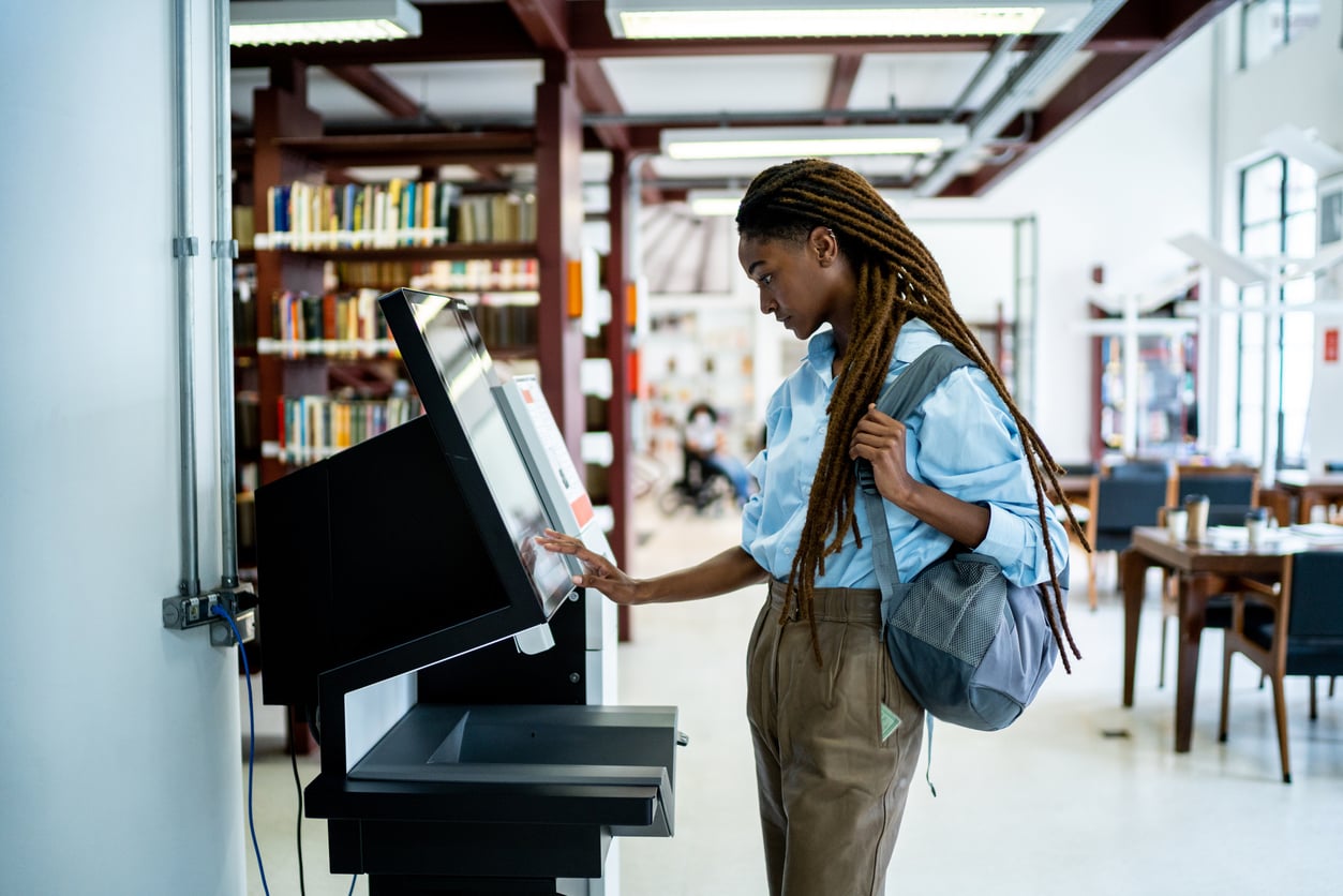 A person with long braids uses a touchscreen kiosk in a library, perhaps exploring resources on money saving habits. They are wearing a blue shirt, brown pants, and a backpack, and appear focused on the screen. Bookshelves and tables are visible in the background.