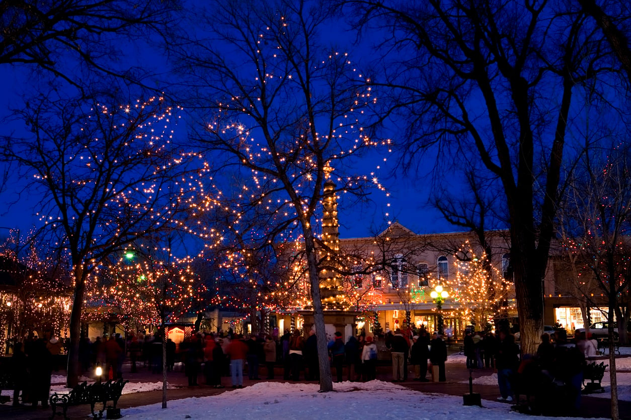 A snowy park at night with trees decorated in glowing holiday lights captures the enchanting spirit found in the best Christmas towns in America. People gather under the lights near a brightly lit building, creating a festive and cheerful winter scene.