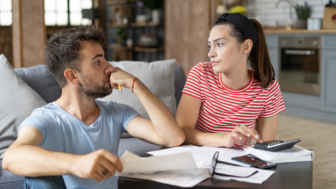 A man and woman sit at a table with papers, a calculator, and a phone, looking serious and thoughtful—possibly discussing whether they’ve spent their budget wisely or fallen for some waste of money products—in a cozy living room setting.