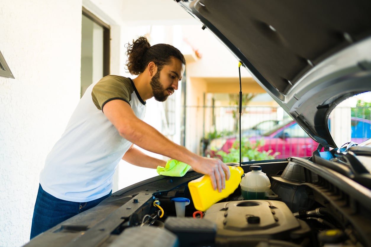 A man with long hair in a ponytail demonstrates car maintenance hacks as he pours fluid from a yellow container into the engine of a car with the hood open, holding a green cloth in his other hand.