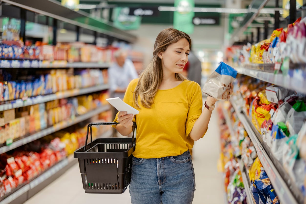 A woman in a yellow shirt holds a shopping basket and a notepad while reading a food package label in a grocery store aisle, evoking the simplicity of life before smartphones.