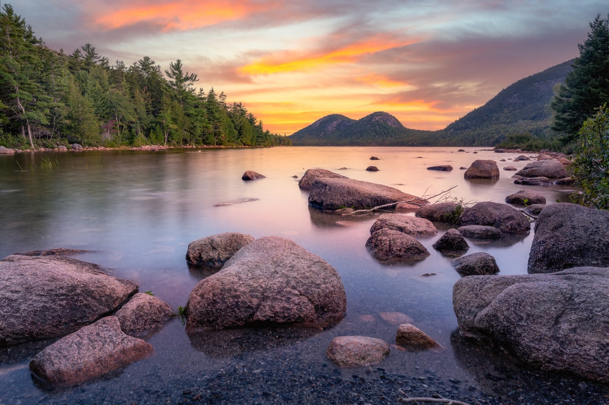 Rocky shoreline of a calm lake at sunset, with orange and purple clouds above pine-covered hills and mountains in the background.