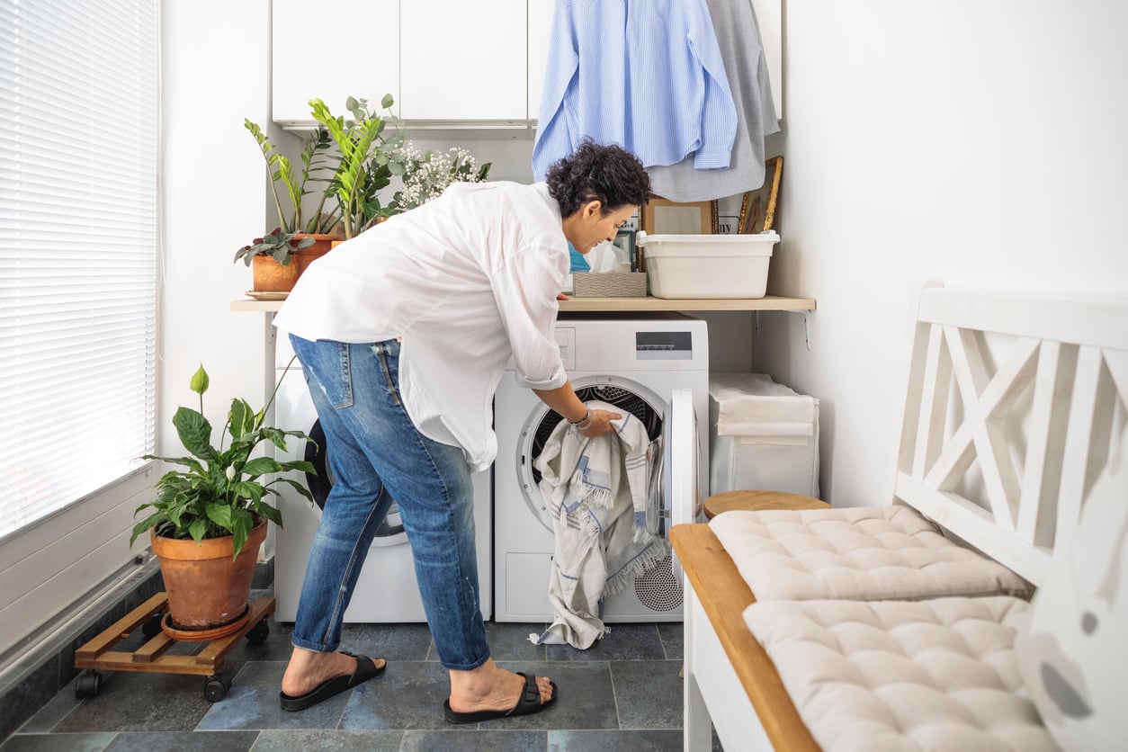 A person with short dark hair, wearing jeans and a white shirt, loads laundry into a washing machine in a bright laundry room with plants and cushioned seating.