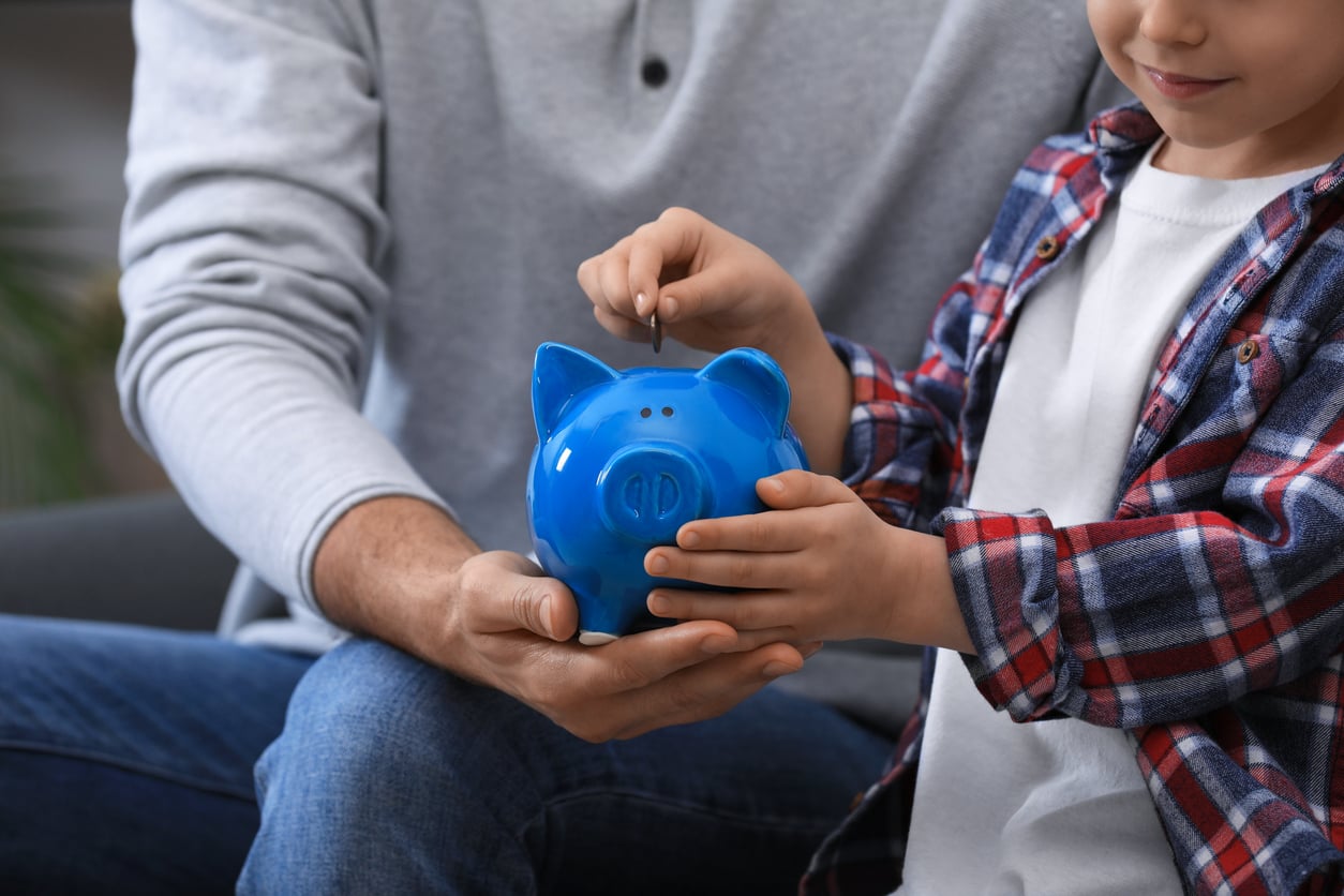 A child and an adult hold a blue piggy bank together as the child puts a coin into it, symbolizing saving money and financial education at a time when families in America are having fewer children.