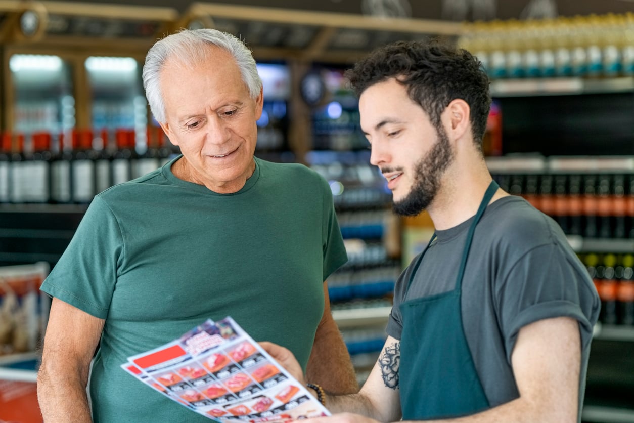 An older man and a young store employee are looking at a colorful flyer together in a grocery store aisle, discussing products or deals.
