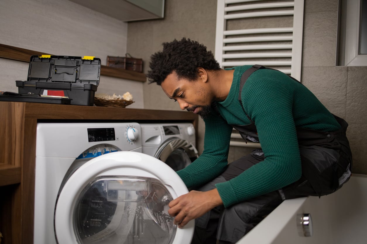 A man in work overalls kneels by a washing machine, inspecting the open door. A toolbox is on the counter behind him, suggesting he is repairing or maintaining the appliance in a modern laundry room—a clear example of practicing money saving habits by handling home repairs himself.