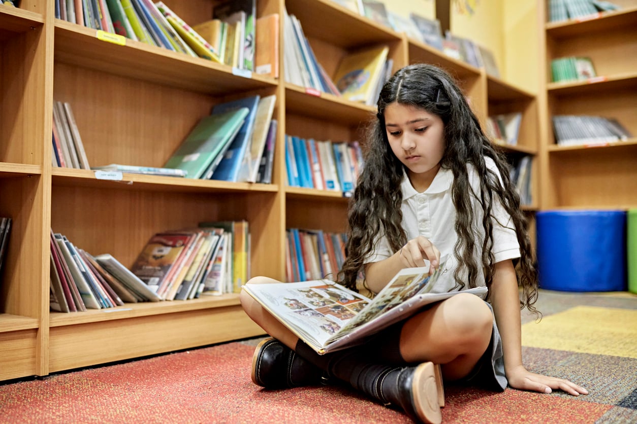 A young girl with long dark hair sits on the floor of a library, reading an illustrated book. She is wearing a white shirt and black shoes. Wooden bookshelves filled with books line the wall behind her.