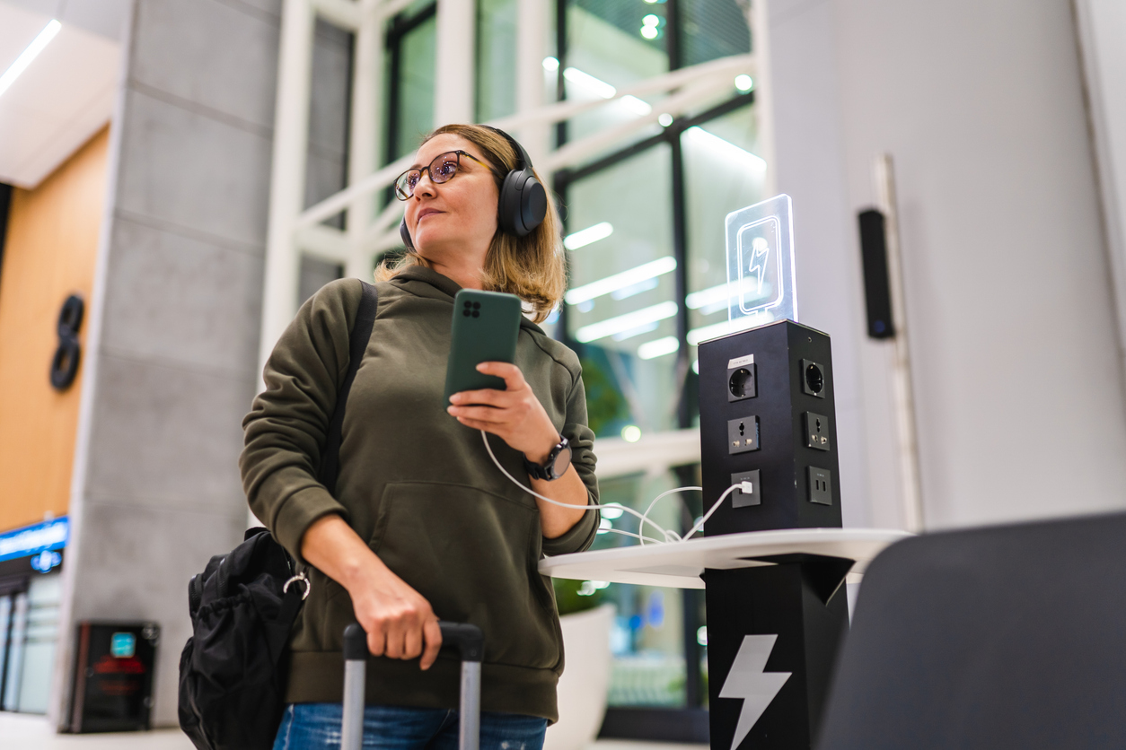 A woman with headphones and glasses stands by a charging station in an airport, holding her phone and suitcase, looking to the side. Modern, brightly lit terminal with large windows in the background.
