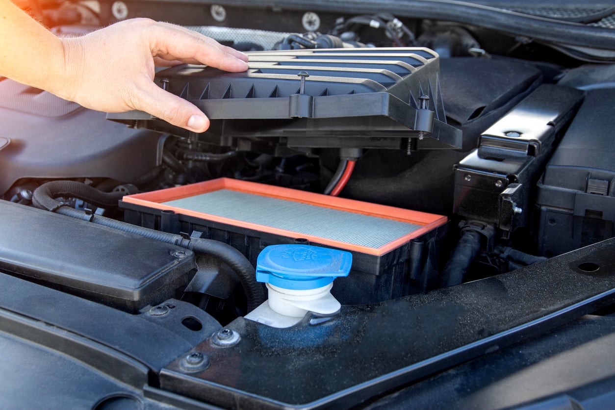 A hand lifts the cover of an air filter housing inside a car engine compartment, revealing a clean rectangular air filter next to a blue windshield washer fluid cap—a simple example of effective car maintenance hacks.