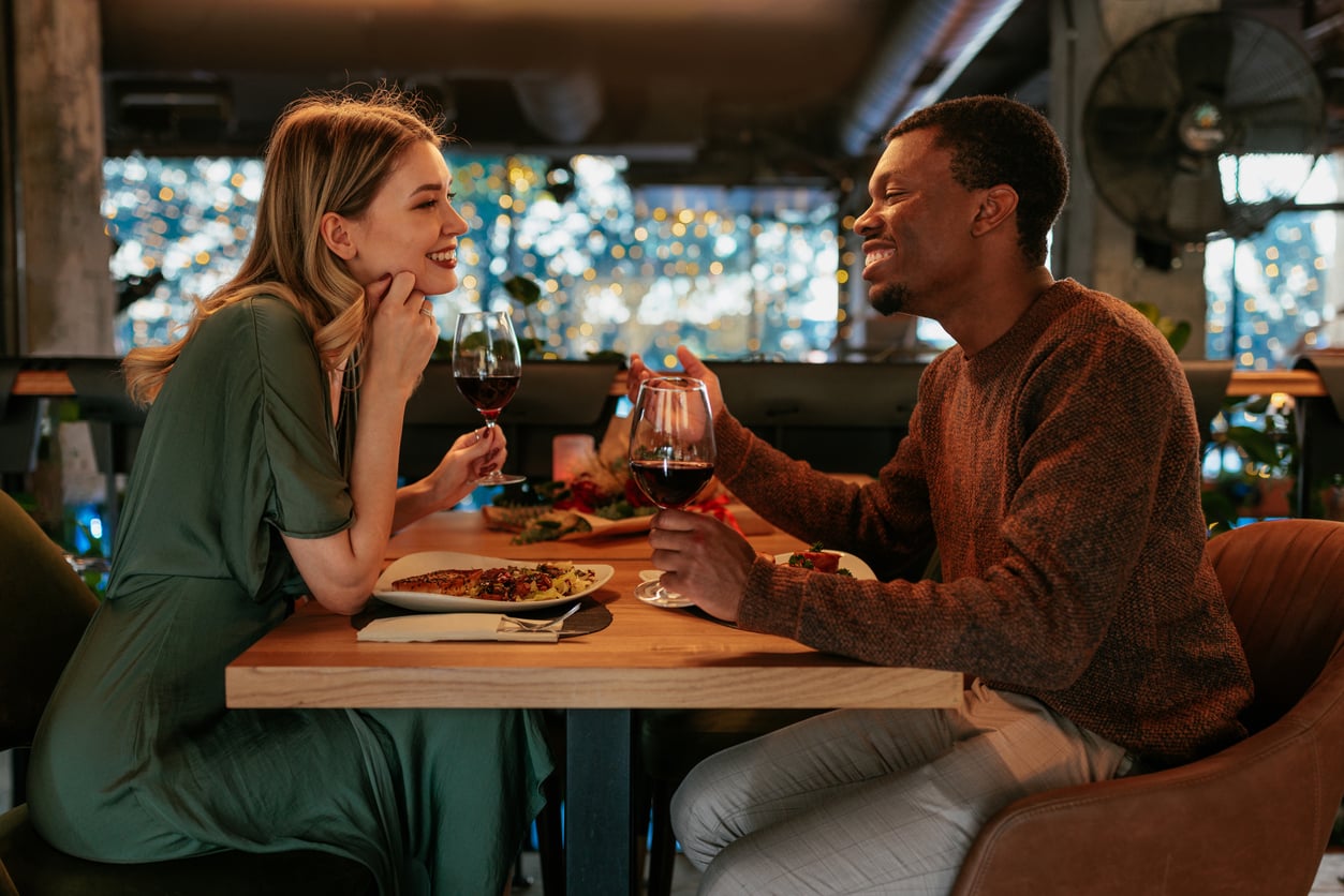 A woman and a man sit across from each other at a restaurant table, smiling and holding glasses of red wine, with plates of food in front of them and string lights in the background.
