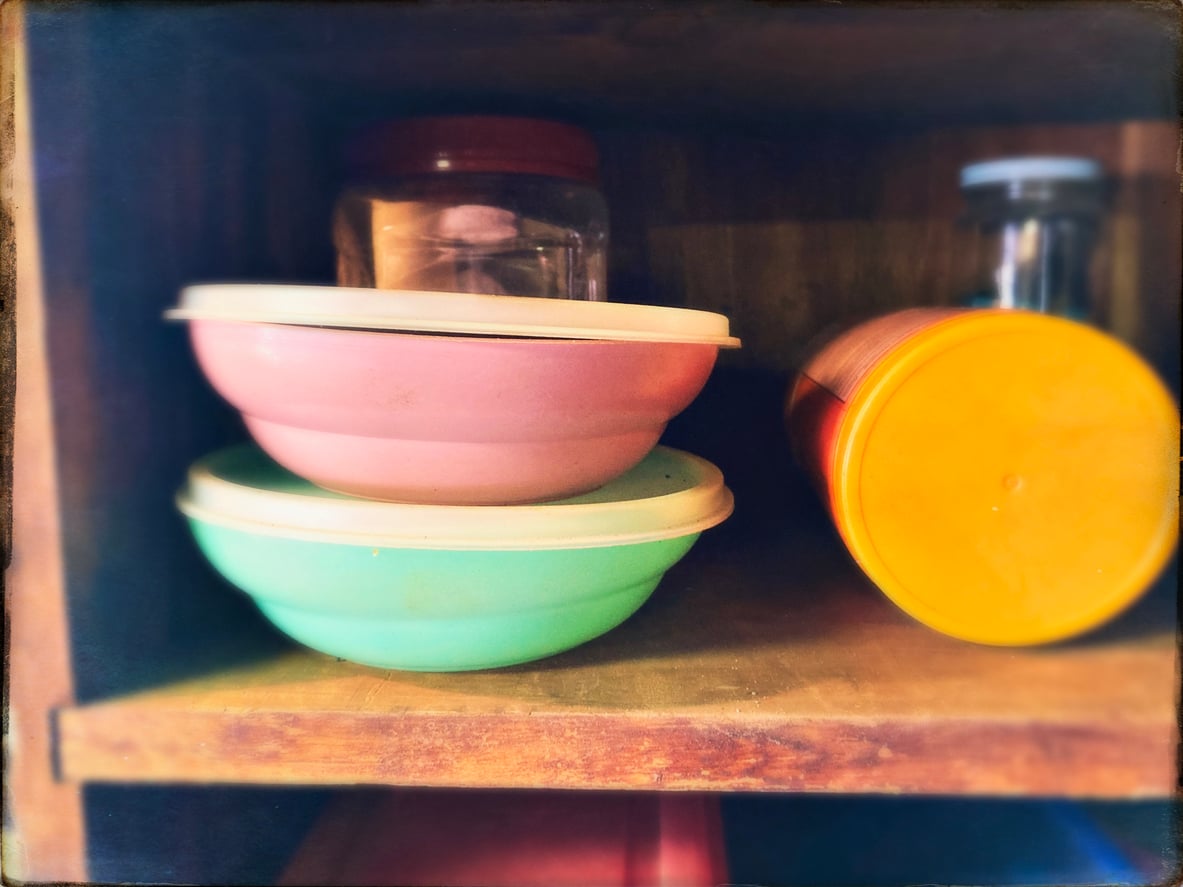 Two plastic bowls, one pink and one light blue, stacked with lids on a wooden shelf among various kitchen items. Beside them are a jar and an orange-lidded container. The scene is slightly blurred at the edges.