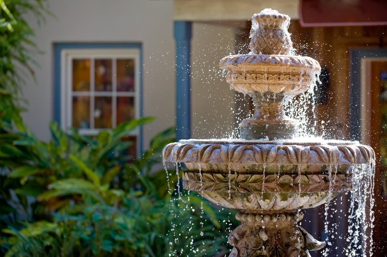 A stone fountain with water cascading over its tiers stands in a sunny garden, surrounded by green plants and thoughtfully arranged outdoor home products. In the blurred background, there is a building with a window and door.