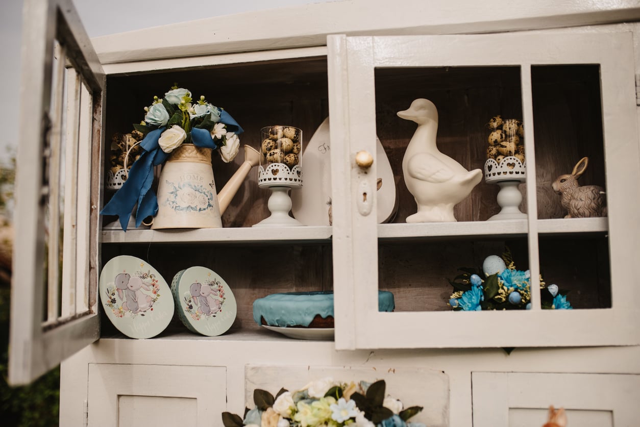 A white cabinet displays decorative plates, a watering can with flowers, a ceramic duck, a small rabbit figure, and jars of quail eggs on stands. Reminiscent of how Middle-Class Moms in the 80s and 90s styled their spaces, blue and white accents create a vintage, spring-themed arrangement.