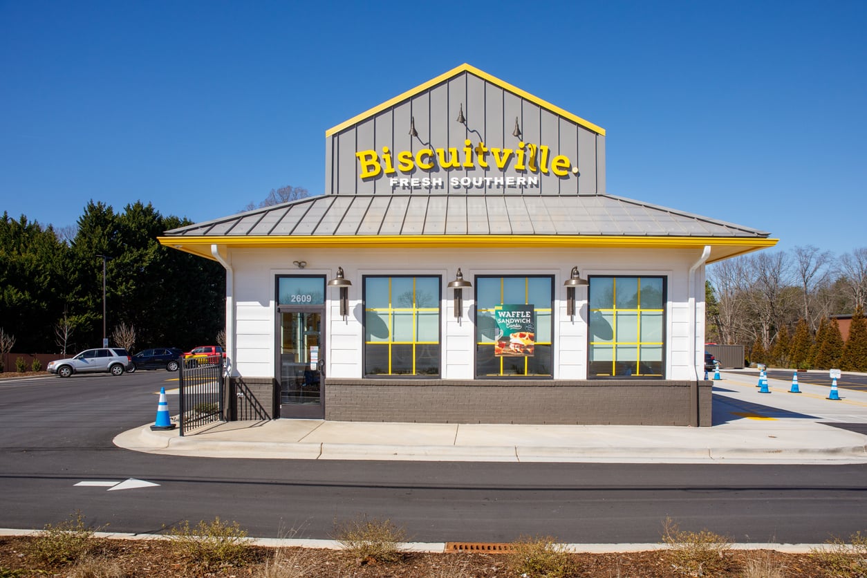 A Biscuitville restaurant, part of the renowned regional fast-food chains, features a gray and yellow roof, large front windows, and a sign that reads “Biscuitville Fresh Southern.” The building is surrounded by a parking lot under a clear blue sky.