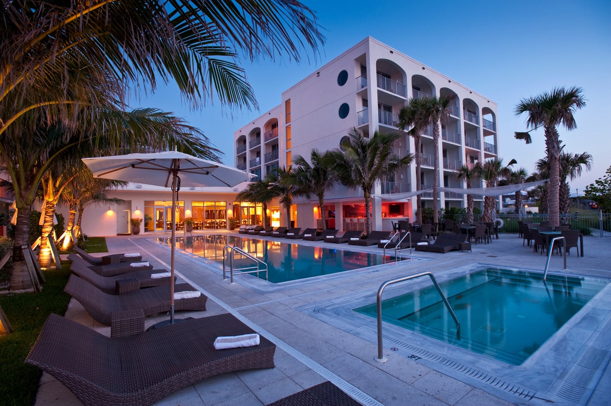 A modern hotel with balconies overlooks a lit outdoor pool area, surrounded by palm trees, lounge chairs, and umbrellas at dusk—a perfect retreat for those seeking relaxation and the best hotel booking tips for a memorable stay.