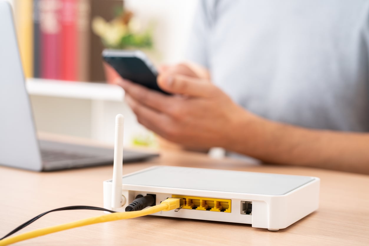 A person managing household expenses on a smartphone sits at a desk, with a white wireless router connected by cables in the foreground. A laptop is partially visible beside the router.